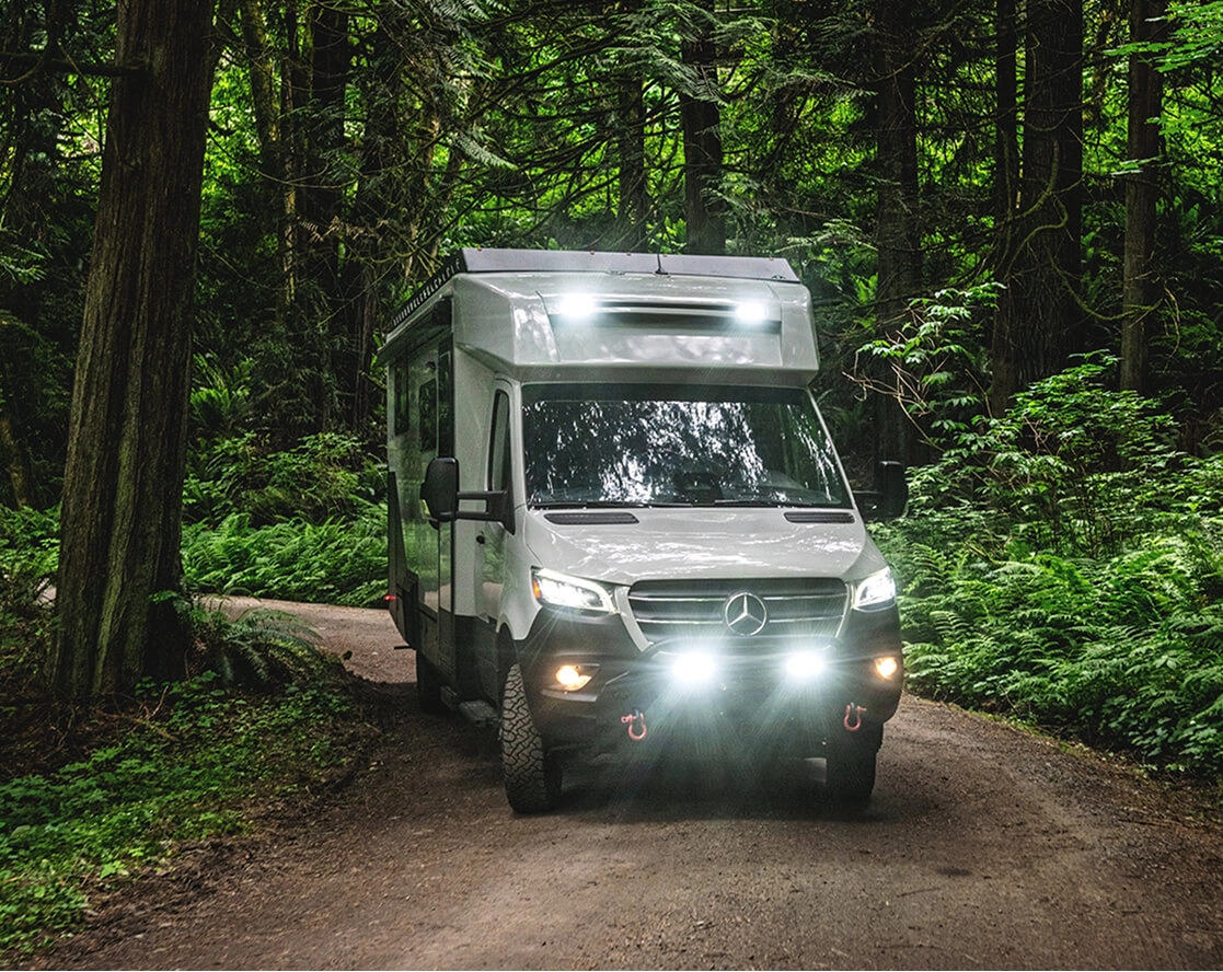 A Tiffin motorhome driving down a wooded path with its lights on